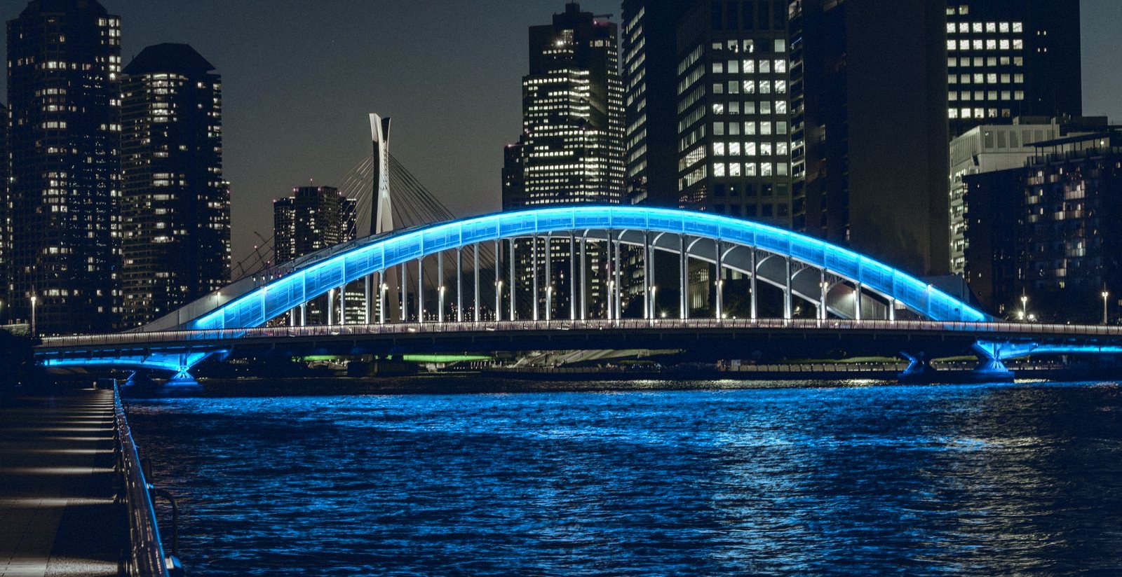 Pont en arc illuminé en bleu au-dessus d'un fleuve, la nuit, devant une ligne de tours éclairées